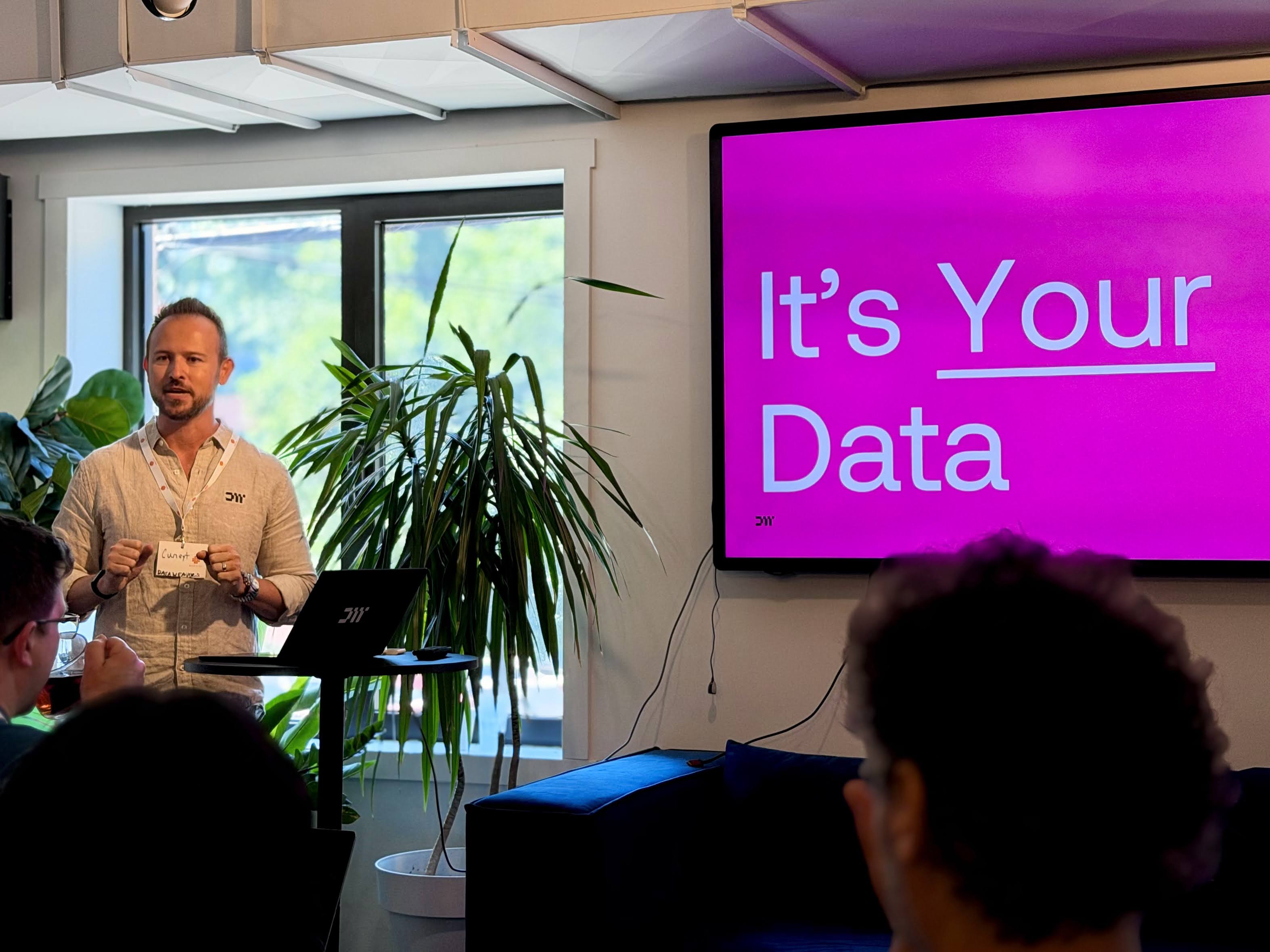 A speaker presents in front of a large screen displaying "It's Your Data," with attendees seated nearby and plants in the background.