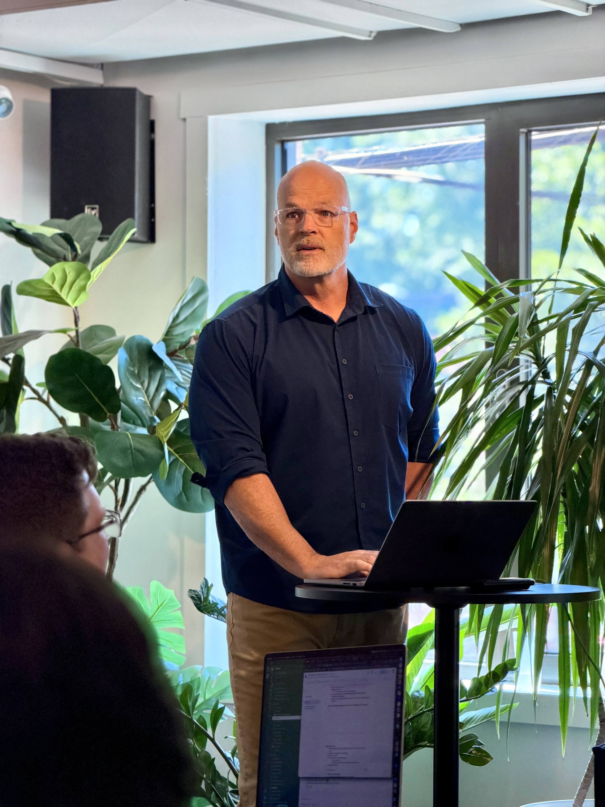 A man with glasses stands at a podium with a laptop, speaking to an audience in a room filled with plants.