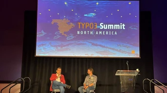 Zwei Redner sitzen auf der Bühne während des TYPO3 Summit Nordamerika, mit einem großen Bildschirm im Hintergrund, der das Veranstaltungslogo und aquatische Visuals zeigt.