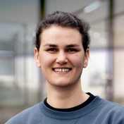 Young woman with dark hair, wearing a gray sweatshirt, smiles confidently in a well-lit indoor setting.