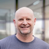 Smiling man with a bald head and beard, wearing a gray shirt, stands in a well-lit indoor space with blurred background elements.