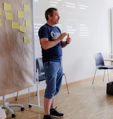 A man in a blue t-shirt stands in front of a presentation screen, gesturing while discussing ideas. Sticky notes are visible on a board nearby.