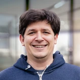 Smiling man with dark hair wearing a blue hoodie, standing in a well-lit environment with blurred background elements.