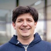 Smiling man with dark hair wearing a blue hoodie, standing in a well-lit environment with blurred background elements.