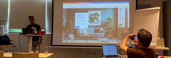 A presenter stands at a podium in a conference room, displaying a digital presentation on a large screen, while an audience member takes photos.
