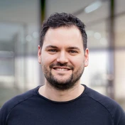 Smiling man with short dark hair and a beard, wearing a black shirt, stands in a modern indoor setting.