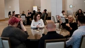 A group of people engaged in discussion around a round table in a conference setting, with additional attendees in the background.