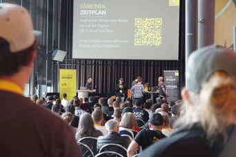 A group of speakers presents on stage at a conference, with an audience seated in front. A QR code and schedule are displayed on a screen.
