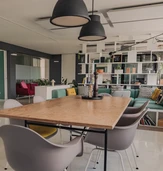 Modern office interior featuring a large wooden table surrounded by gray chairs, colorful seating areas, and a bookshelf with various items.