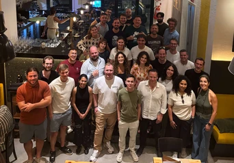 A large group of people poses together in a restaurant, smiling and standing in two rows, with a bar visible in the background.
