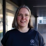 Person mit kurzen, glatten Haaren, die eine Brille trägt und ein schwarzes T-Shirt mit einem Logo anhat. Der Hintergrund ist neutral und unauffällig.