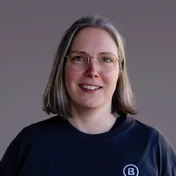 Person mit kurzen, glatten Haaren, die eine Brille trägt und ein schwarzes T-Shirt mit einem Logo anhat. Der Hintergrund ist neutral und unauffällig.