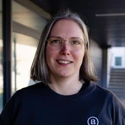 Person mit kurzen, glatten Haaren, die eine Brille trägt und ein schwarzes T-Shirt mit einem Logo anhat. Der Hintergrund ist neutral und unauffällig.