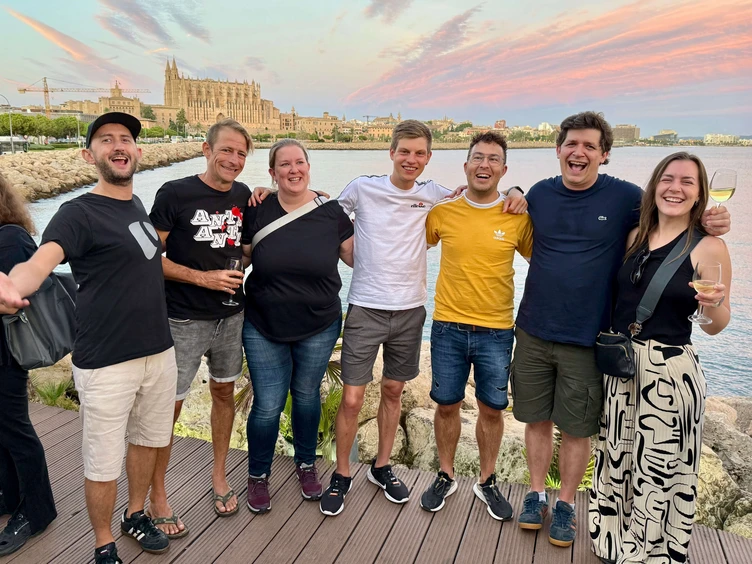 A group of seven people stands together by the waterfront, smiling and posing for the photo. They are casually dressed, with some holding drinks, and the backdrop features the cityscape of La Palma, Mallorca in front of a colorful sky.
