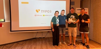 Four individuals stand in front of a presentation screen displaying the TYPO3 logo and sponsor logos, holding an award or certificate.