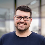 Smiling man with short brown hair and glasses, wearing a navy blue shirt, stands in a modern indoor setting.