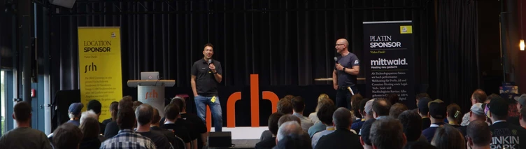 Two speakers present on stage at a conference, with an audience seated in front. A slide displays information about an event in 2027.