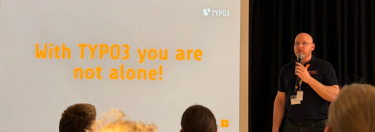 Ein Referent präsentiert auf einer Konferenz, mit einer Folie, die in fettem Orange den Text „Mit TYPO3 sind Sie nicht allein!“ zeigt.