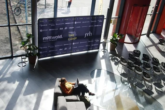 A person sits on a sofa working on a laptop in a modern lobby with a large promotional backdrop and empty chairs.