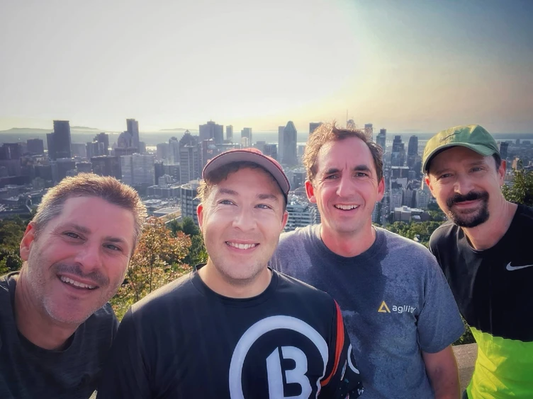 Four men pose for a selfie with a city skyline in the background, showcasing a sunny day and urban landscape.