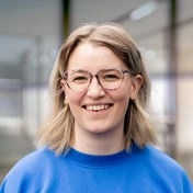Smiling woman with shoulder-length blonde hair, wearing glasses and a blue sweatshirt, stands in a bright, modern environment.