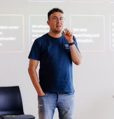 A man in a navy t-shirt and shorts gestures while speaking in front of a presentation screen with text in German.