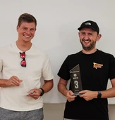 Zwei Männer stehen nebeneinander, einer hält einen Pokal mit der Aufschrift "3" und der andere hält einen kleinen Gegenstand, beide lächeln.