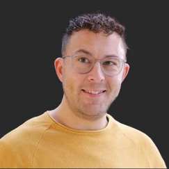 A man with short, curly hair and glasses smiles while wearing a yellow sweater. The background is dark, emphasizing his features.