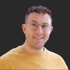 A man with short, curly hair and glasses smiles while wearing a yellow sweater. The background is dark, emphasizing his features.