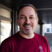 Lächelnder Mann mit kurzen Haaren und einem Bart, der ein weinrotes Hemd mit einem Logo trägt, vor einem neutralen Hintergrund.