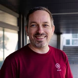 Lächelnder Mann mit kurzen Haaren und einem Bart, der ein weinrotes Hemd mit einem Logo trägt, vor einem neutralen Hintergrund.
