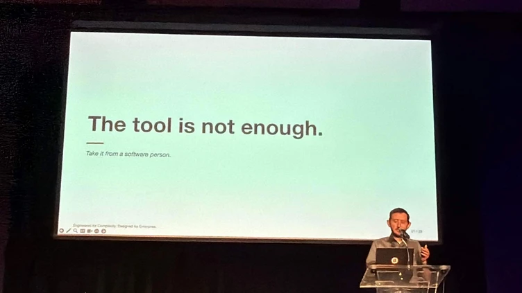 Ein Redner steht an einem Podium vor einer Präsentationsfolie, auf der steht: "Das Werkzeug ist nicht genug. Nehmen Sie es von einer Software-Person.