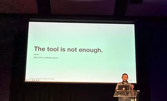 A speaker stands at a podium in front of a presentation slide that reads, "The tool is not enough. Take it from a software person.