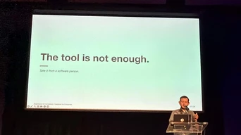Ein Redner steht an einem Podium vor einer Präsentationsfolie, auf der steht: "Das Werkzeug ist nicht genug. Nehmen Sie es von einer Software-Person.