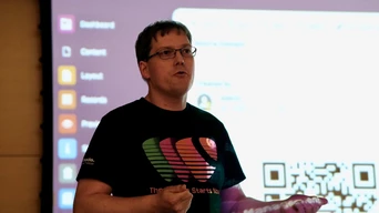 A speaker in a black t-shirt gestures while presenting in front of a screen displaying a dashboard interface and QR code.