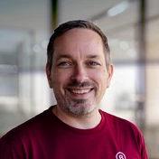 Smiling man with short hair and a beard, wearing a red t-shirt, stands in a well-lit indoor space with blurred background.
