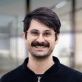 Mann mit Brille und Schnurrbart, der einen schwarzen Pullover trägt und in einer modernen Innenumgebung lächelt.
