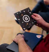 A person holds a colorful booklet featuring a circular logo with a "B," sitting next to a laptop on their lap.