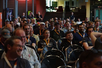 Ein diverses Publikum von Menschen, die in einem Konferenzsaal sitzen, lächeln und engagiert sind, wobei einige Namensschilder und Lanyards tragen.
