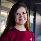 Junge Frau mit langen braunen Haaren, die ein kastanienbraunes Shirt trägt und vor einem neutralen Hintergrund lächelt.