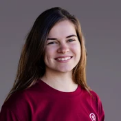 Junge Frau mit langen braunen Haaren, die ein kastanienbraunes Shirt trägt und vor einem neutralen Hintergrund lächelt.