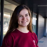 Young woman with long brown hair, wearing a maroon shirt, smiling against a neutral background.