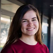 Young woman with long brown hair, wearing a maroon shirt, smiling against a neutral background.