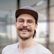 Man with a mustache wearing glasses and a cap, smiling at the camera. He is dressed in a light-colored shirt, with a blurred background.