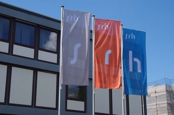Three flags displaying the letters "s," "r," and "h" in white on colored backgrounds, positioned outside a modern building.