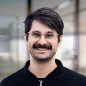Man with glasses and a mustache, wearing a black sweater, smiling in a modern indoor setting.