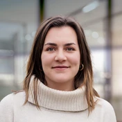 Young woman with long, wavy hair wearing a light sweater, smiling softly against a blurred indoor background.