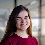 Young woman with long brown hair, wearing a maroon shirt, smiles confidently in a well-lit indoor setting.