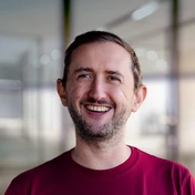 Smiling man with short hair and a beard, wearing a maroon shirt, stands in a modern indoor setting with blurred background elements.