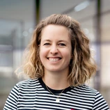 Smiling woman with curly hair wearing a striped shirt and necklace, standing in a bright indoor setting.